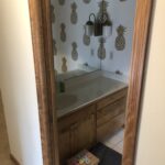 The original, dated powder room featuring distinctive gold pineapple wallpaper, a simple oak vanity, and a large mirror with a light fixture mounted on it, a before photo of a powder room remodel in Edina, MN.