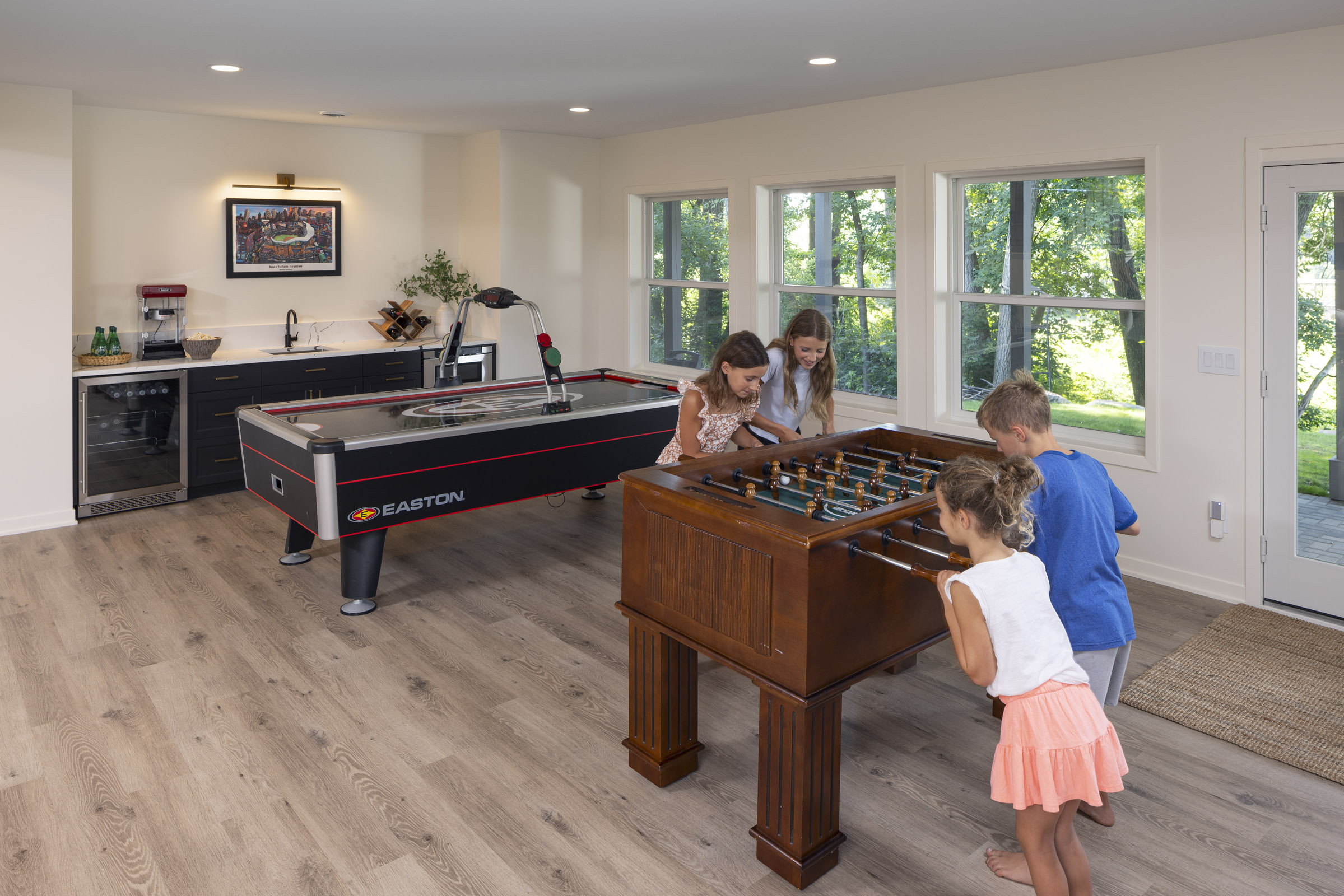 A fun and lively lower level game room, with children gathered around the foosball table for a game, and an air hockey table and wet bar in the background, part of a lower level remodel in Edina, MN.