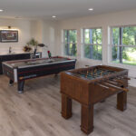 A bright and spacious lower level game room, featuring both a foosball table and an air hockey table, a custom wet bar, and large windows with a view to the backyard, part of a lower level remodel in Edina, MN.
