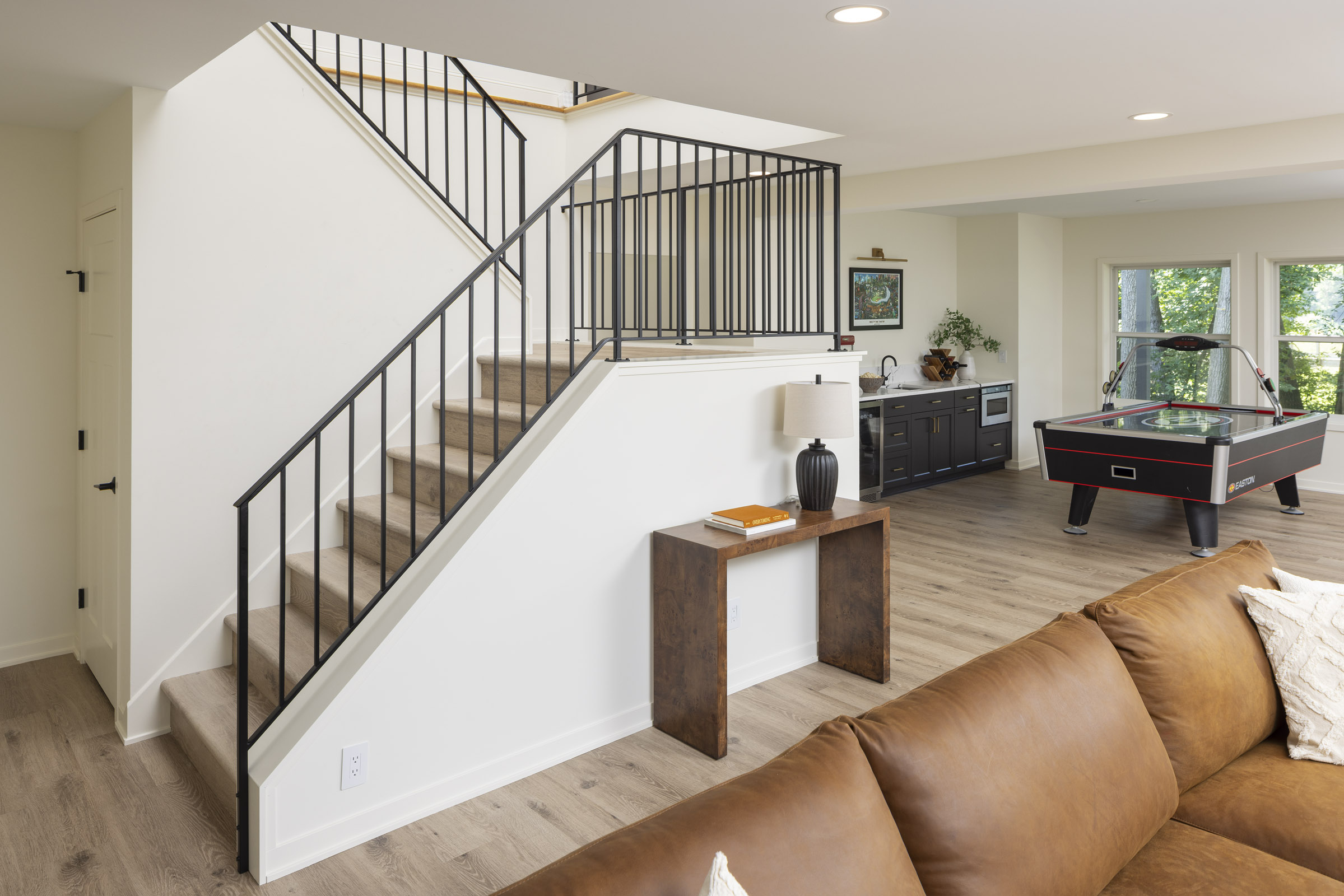 A modern open staircase with a simple black metal railing that descends into the lower level, with a view of the game room's air hockey table and wet bar in the background, part of a lower level remodel in Edina, MN.