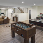 A spacious, multi-zone lower level designed for entertainment, featuring a game area with foosball and air hockey tables, a custom wet bar, a central lounge, and a glass-enclosed home gym, part of a lower level remodel in Edina, MN.
