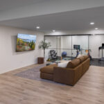 A wide view of a modern lower level, showcasing the comfortable lounge with its leather sectional, the adjacent glass-enclosed home gym, and a view into the nearby bathroom, part of a lower level remodel in Edina, MN.