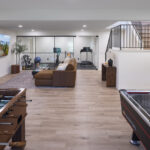 A wide view of a spacious and multi-functional remodeled lower level, showcasing a game room with foosball and air hockey, a central lounge area, and a glass-enclosed home gym in the background, part of a lower level remodel in Edina, MN.