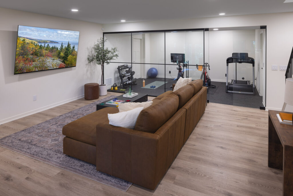 A modern, multi-functional lower level featuring a comfortable lounge with a large leather sectional and a wall-mounted TV, with a full view into the adjacent glass-enclosed home gym, part of a lower level remodel in Edina, MN.