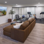 A modern, multi-functional lower level featuring a comfortable lounge with a large leather sectional and a wall-mounted TV, with a full view into the adjacent glass-enclosed home gym, part of a lower level remodel in Edina, MN.