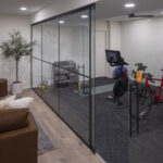 A modern lower level featuring a custom, glass-enclosed home gym with rubber flooring, adjacent to a comfortable lounge area with light wood-look floors, part of a lower level remodel in Edina, MN.
