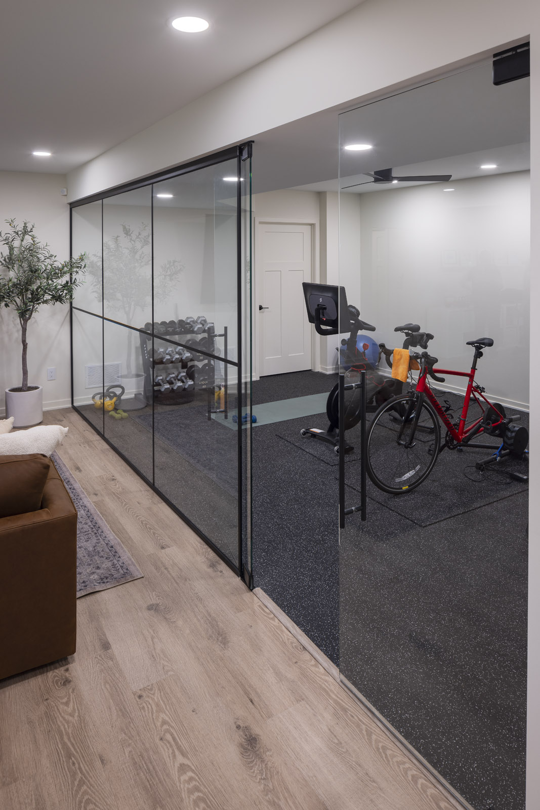 A modern lower level featuring a custom, glass-enclosed home gym with rubber flooring, adjacent to a comfortable lounge area with light wood-look floors, part of a lower level remodel in Edina, MN.