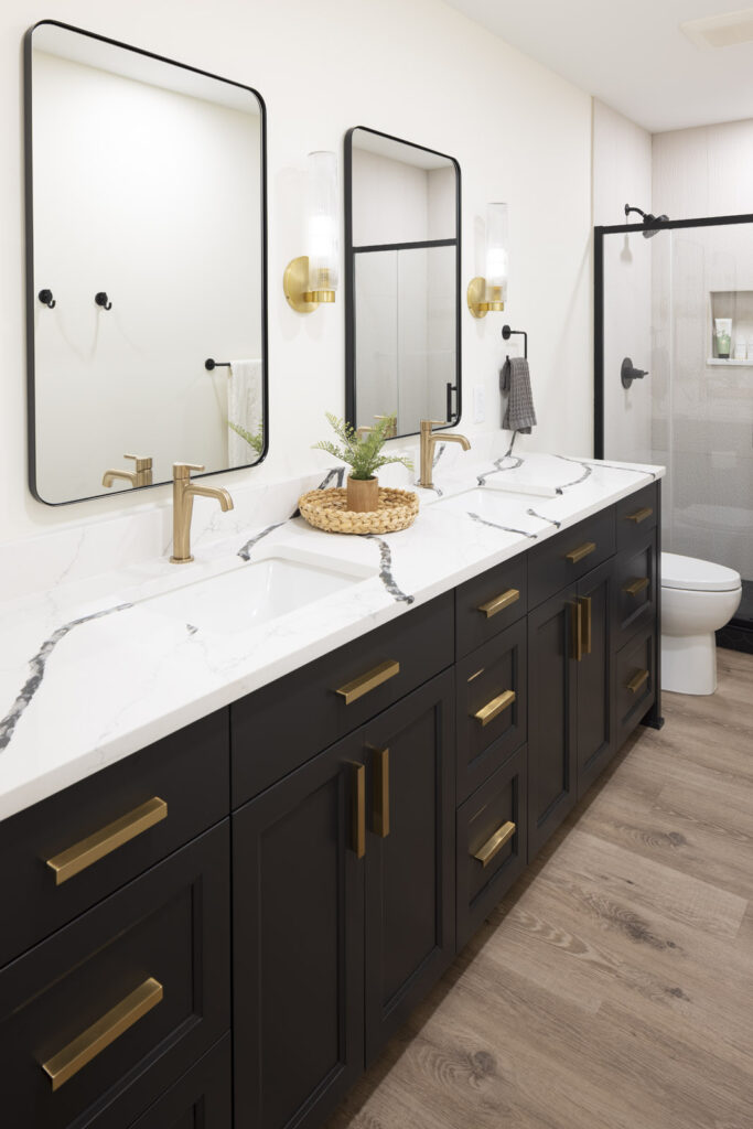 A striking modern bathroom featuring a long, dark double vanity topped with white quartz with dramatic black veining, and accented with black-framed mirrors and brushed brass fixtures, part of a lower level remodel in Edina, MN.