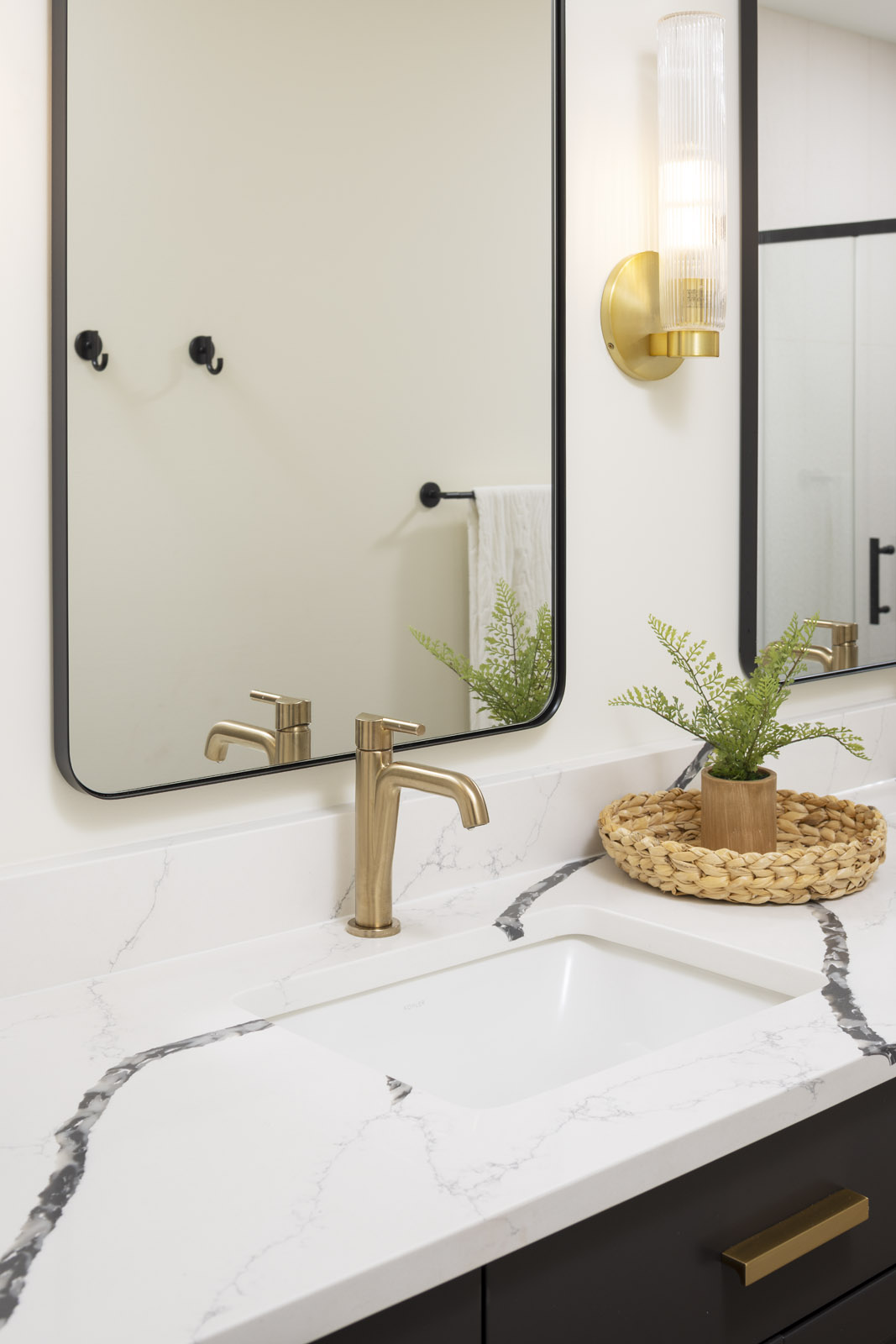 A detailed view of a modern bathroom vanity, featuring a white quartz countertop with dramatic black veining, a brushed brass faucet, and a modern brass and glass wall sconce, part of a lower level remodel in Edina, MN.
