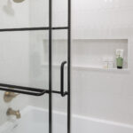 A modern and stylish tub and shower combination featuring a black-framed sliding glass door, glossy white scalloped wall tiles, and brushed gold fixtures, part of a lower level remodel in Edina, MN.