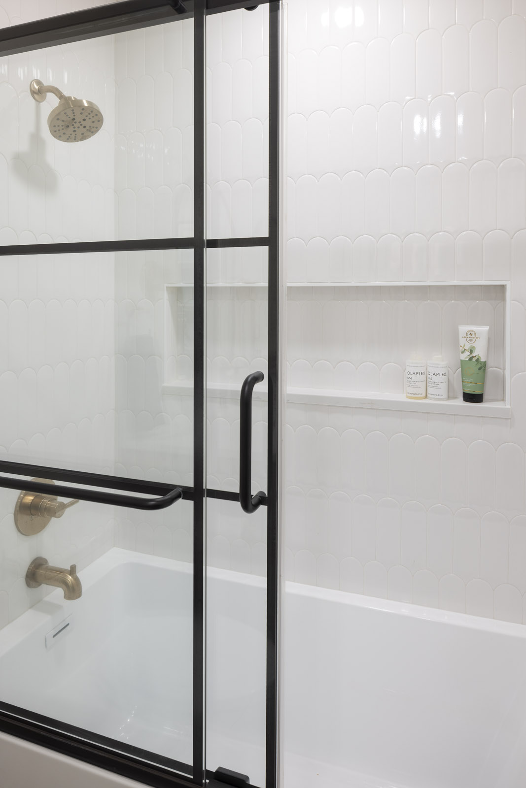 A modern and stylish tub and shower combination featuring a black-framed sliding glass door, glossy white scalloped wall tiles, and brushed gold fixtures, part of a lower level remodel in Edina, MN.