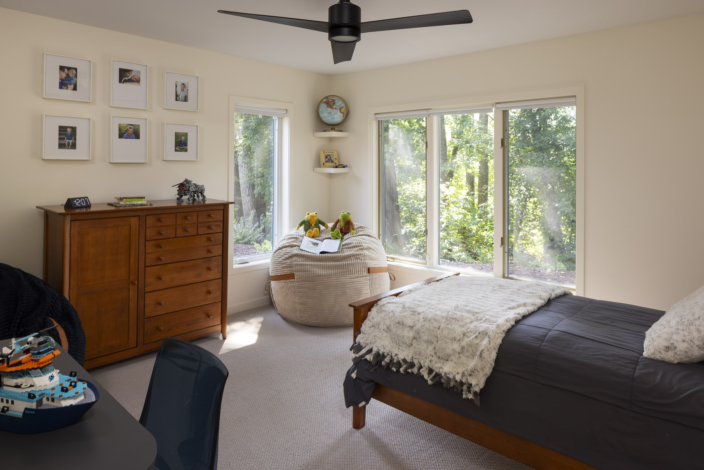 A bright and comfortable kid's bedroom featuring a cozy reading nook with a large beanbag chair, a traditional wood dresser, and large windows with a view of the wooded backyard, part of a lower level remodel in Edina, MN.